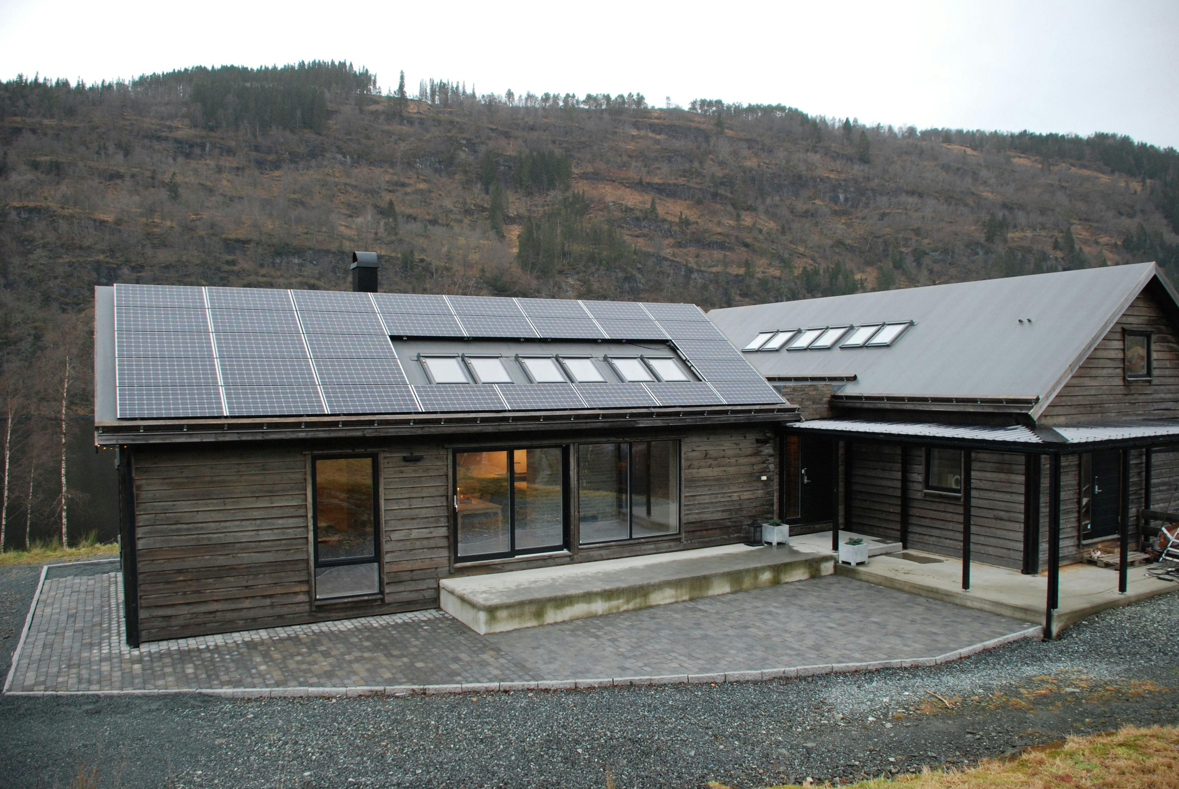 Wooden house with solar panels and a porch next in front of a hill with pine trees
