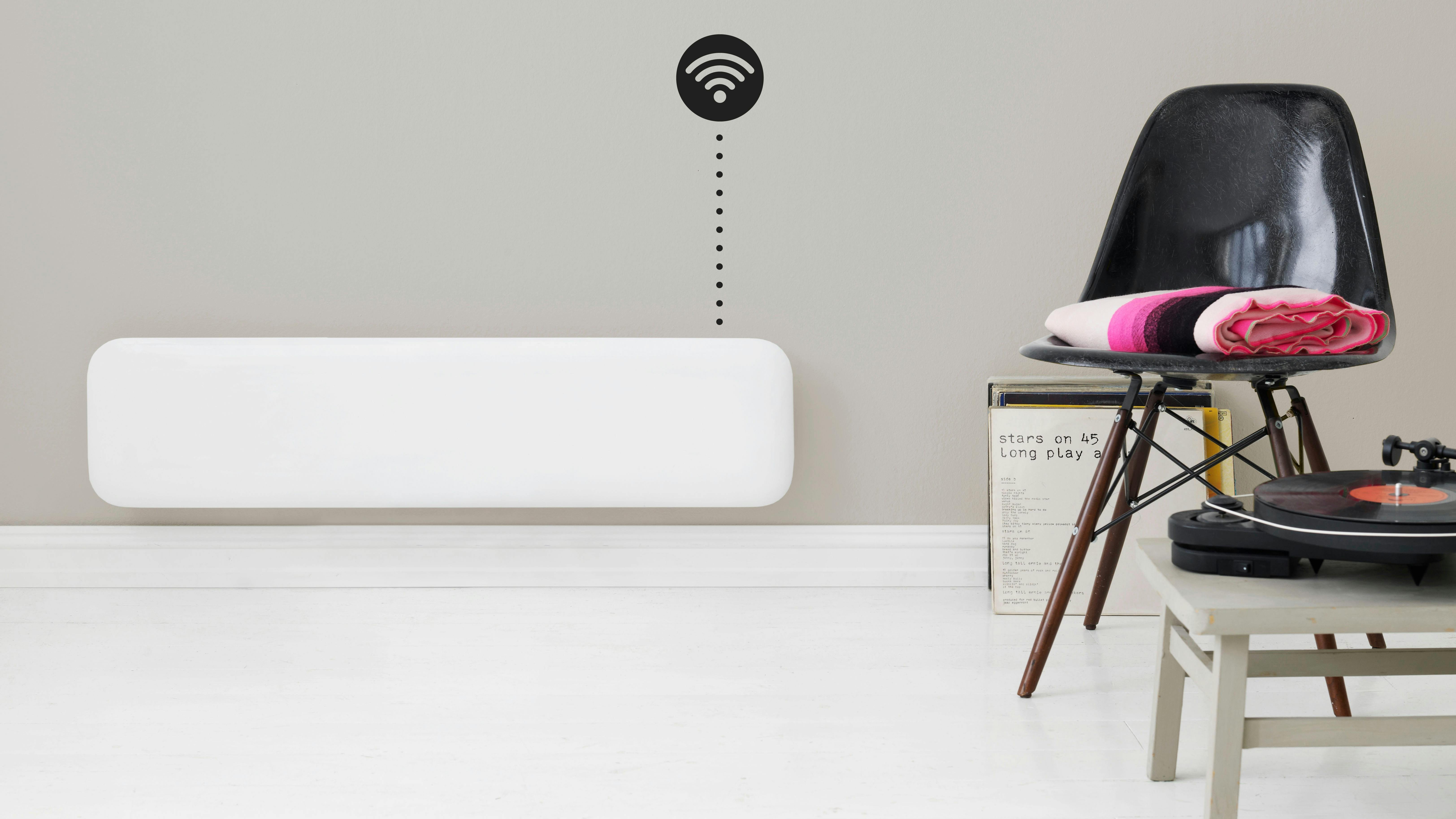 Mill panel heater mounted to the wall with next to it some vinyl records, a black chair with a folded blanket and a side table with a record player on it
