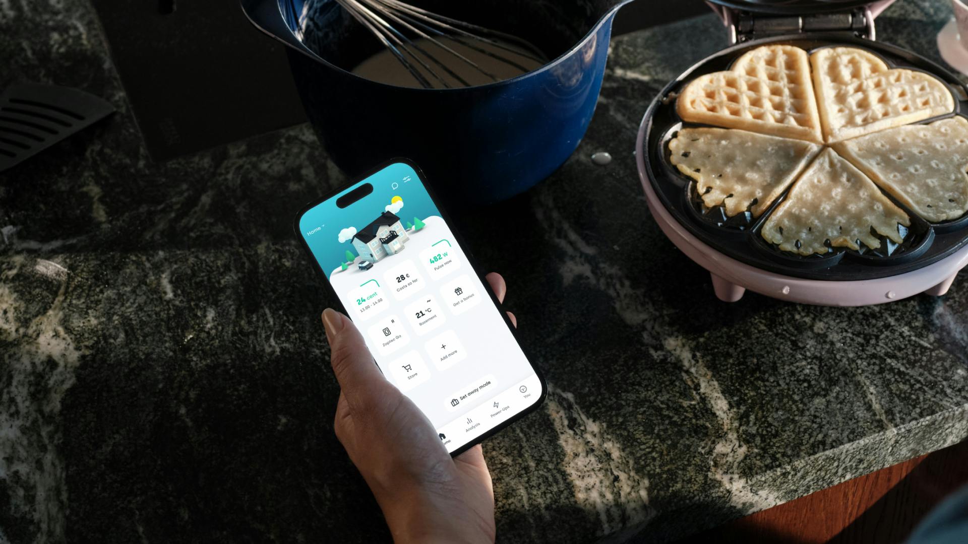 Woman holding a phone showing the smart heating screen in de Tibber app with a picture frame and vase in the background